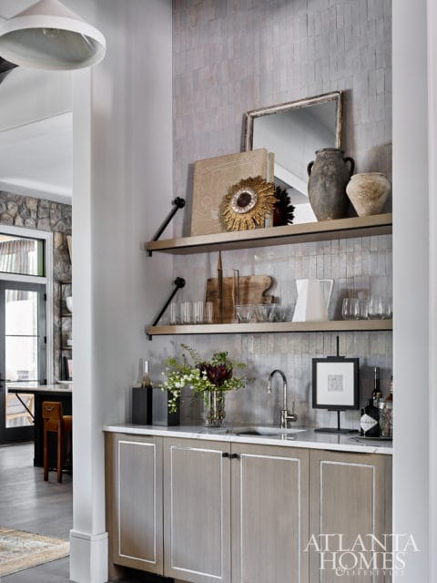 Bar area with light wood cabinetry, marble countertop, and a stainless steel faucet. Shelves above display decorative items and glassware.