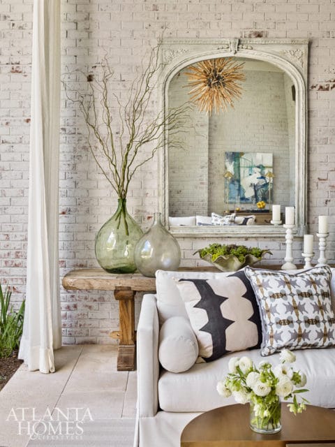 White sofa with patterned pillows in front of a rustic wooden console table topped with glass vases and dried branches. A large arched mirror hangs above the table.