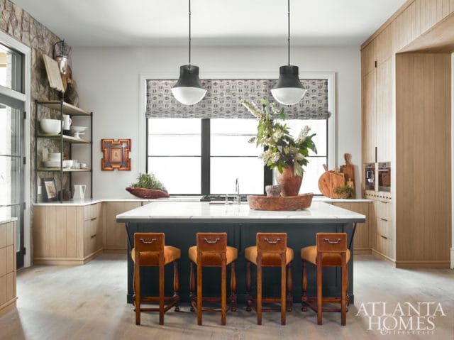 Dark green kitchen island with marble countertop, four orange velvet bar stools, and a large window with a patterned shade.