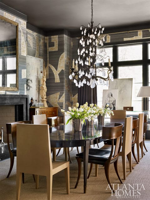 Dining room with a dark wood table set with white tulips, surrounded by chairs of various wood tones and upholstered seats, under a crystal chandelier.