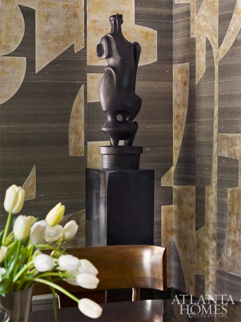Dark abstract sculpture on a black pedestal against a textured wallpaper with geometric patterns in beige and gray. A bouquet of white tulips is in the foreground.