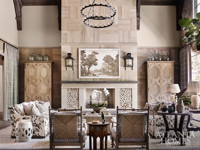 Living room with a stone fireplace, wood paneled walls, two upholstered sofas, two woven chairs, and a circular chandelier.