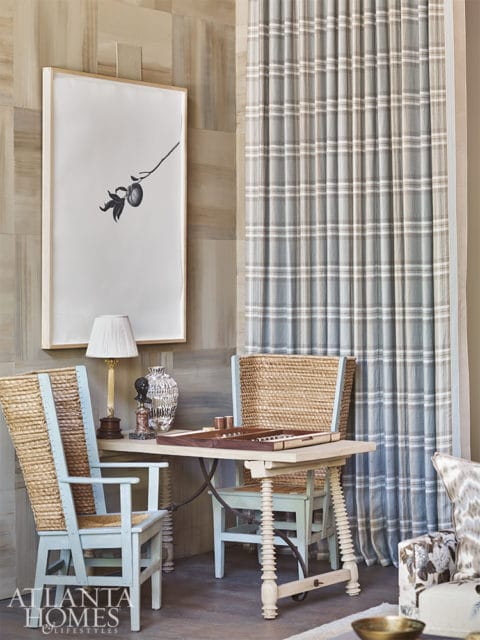 Light blue chairs with woven seats and backs flank a table with a backgammon set, next to plaid curtains and a framed botanical print on a wood-paneled wall.