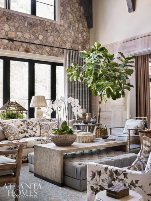 Living room with stone accent wall, floral upholstered sofa and chairs, tufted ottoman, and large fiddle leaf fig plant.
