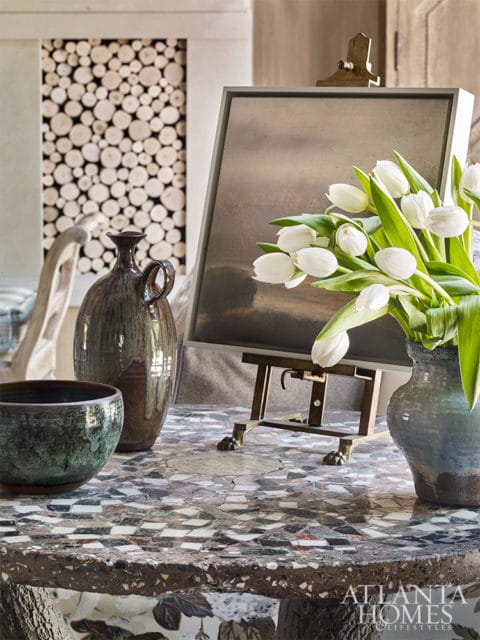 Interior scene with a mosaic-topped table featuring a dark green ceramic bowl, a tall brown glazed vase, white tulips in a blue vase, and a framed artwork on an easel.