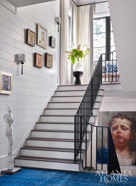 White shiplap walls with a gallery of portraits, a blue rug, and a staircase with black railings leading upstairs. A large painting of a child's face is on the right wall.