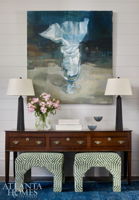 Entryway console table with two green geometric upholstered ottomans, a vase of pink peonies, two black lamps, and abstract art of a glass vase.