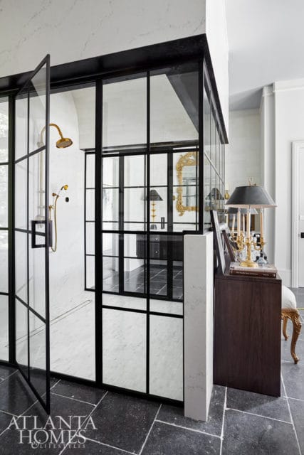 Modern bathroom with black-framed glass shower enclosure, marble walls, brass shower fixtures, dark wood vanity, and ornate gold mirror.