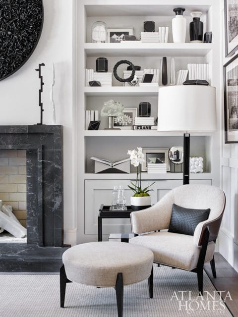 Living room with a marble fireplace, built-in white shelves filled with black and white decor, a cream armchair with a black pillow, and an ottoman.