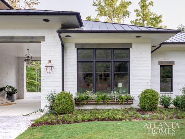 Modern white brick house with black metal roof and windows, featuring a covered porch with hanging lanterns and a window box garden.