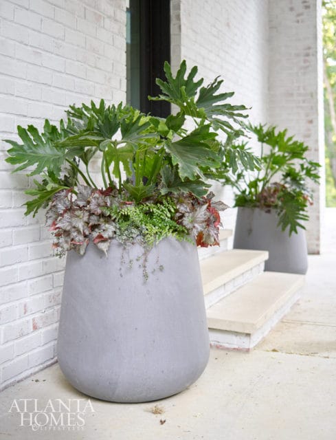 Two large gray planters filled with lush green plants and smaller red-leafed plants sit on a porch next to light-colored stone steps.