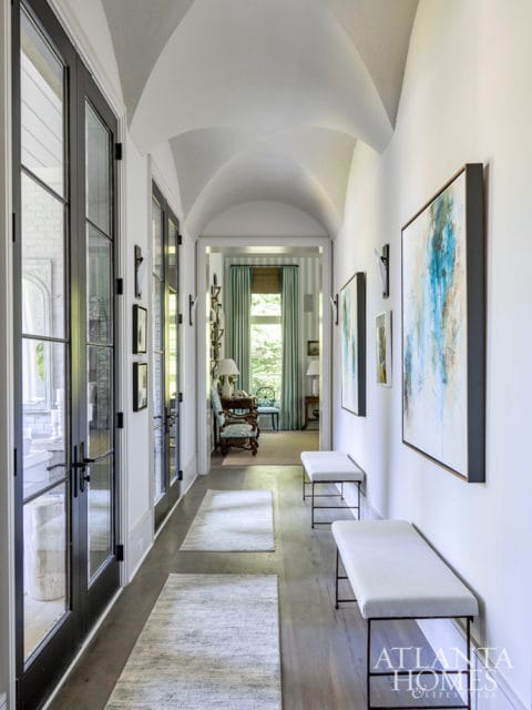 Hallway with vaulted ceiling, black-framed glass doors, abstract art, and white benches. Rugs line the wooden floor.