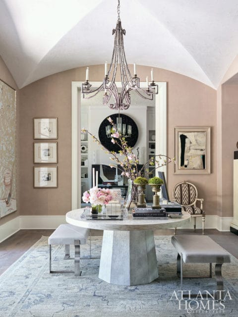 Foyer with a pale pink vaulted ceiling, a faceted pedestal dining table with benches, and a crystal chandelier. A decorative rug covers the floor.