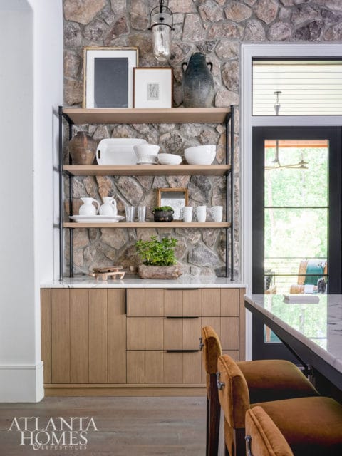 Built-in shelving unit with natural stone backsplash, displaying various ceramic and glass decorative items, above light wood cabinetry with black hardware.