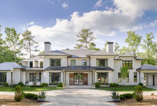 Modern white brick house with black trim windows, dark metal roof sections, and two prominent brick chimneys. A paved driveway leads to the double wooden front doors.
