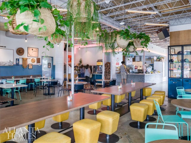 Restaurant interior with wood tables, yellow square stools, and teal chairs. Hanging plants and neon lighting add to the decor.