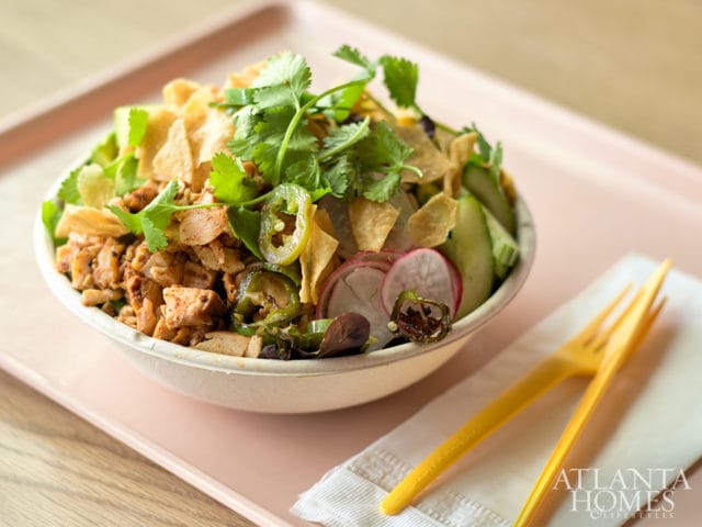 Chicken salad bowl with cilantro, jalapeños, radishes, cucumbers, and tortilla chips, served with a yellow fork on a pink tray.