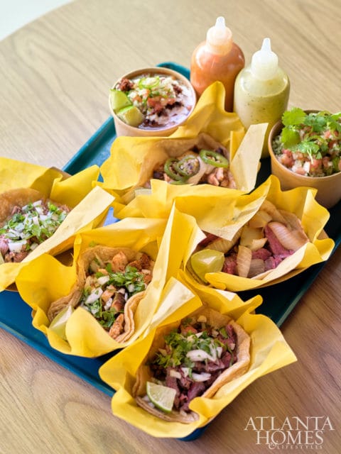 Assortment of tacos with various fillings, served in yellow paper wrappers on a blue tray, with small cups of salsa and two squeeze bottles of sauce.