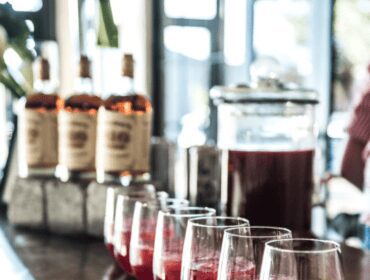 Row of stemless glasses filled with red liquid and ice, behind them are bottles of liquor.