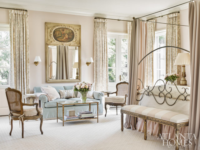 Accented with draperies and luxurious bedding, the light-filled master bedroom boasts a seating area with a plush sofa in light blue fabric and antique chairs. The sconces are from Oliver Street Designs.