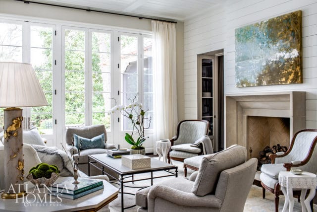 Living room with a stone fireplace, large windows overlooking greenery, and upholstered seating. Abstract art hangs above the fireplace.