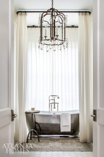 Freestanding silver bathtub with chrome fixtures, white towels, and a crystal chandelier above. Sheer white curtains frame the window behind the tub.