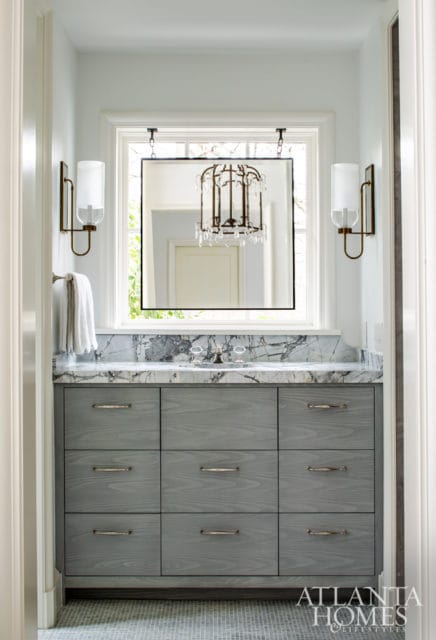 Bathroom vanity with gray wood drawers and a marble countertop. A large mirror hangs above the sink, reflecting a chandelier.