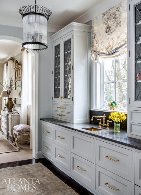 Light gray kitchen cabinets with brass hardware and dark gray countertops, featuring a brass sink and faucet under a floral Roman shade window.