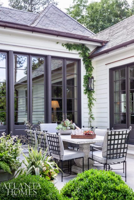 Patio dining set with stone table and chairs surrounded by lush greenery, featuring large dark-framed windows and a climbing vine on a white clapboard house.