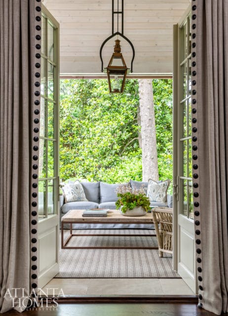 Covered porch with light blue sofa, wooden coffee table, and wicker chair, overlooking lush green trees. Atlanta Homes & Lifestyles logo in bottom left.