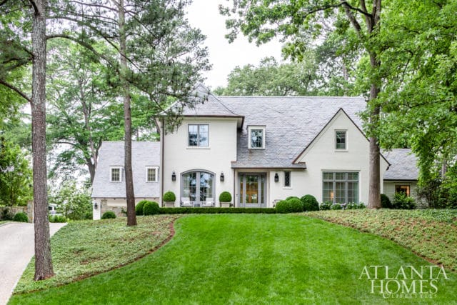 White stucco house with gray slate roof, arched front door, large windows, and manicured landscaping with lush green lawn and mature trees.