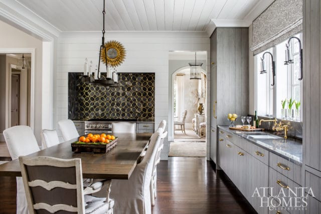 Dining area with dark wood table and white upholstered chairs, adjacent to a kitchen with gray cabinetry, marble countertops, and a gold-accented backsplash.