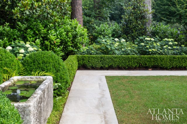 Stone water trough with lily pads, manicured boxwood hedges, hydrangeas, and a stone pathway in a lush garden