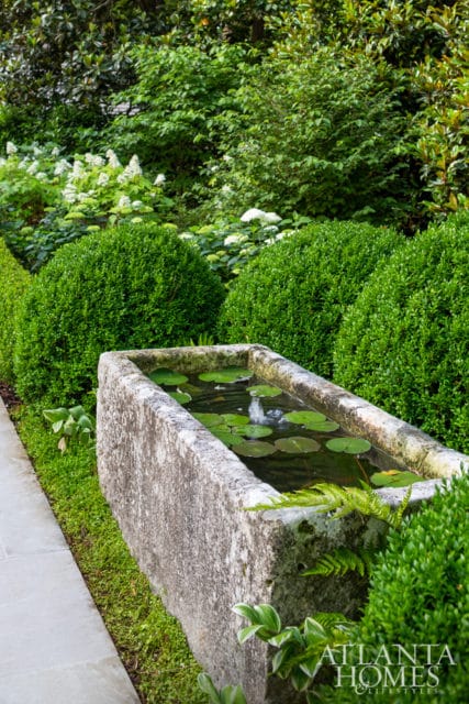 Stone trough water feature with lily pads and a fountain, surrounded by manicured boxwood hedges and lush greenery.