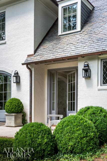 White brick house exterior with slate roof, gray double doors, and manicured boxwood bushes.