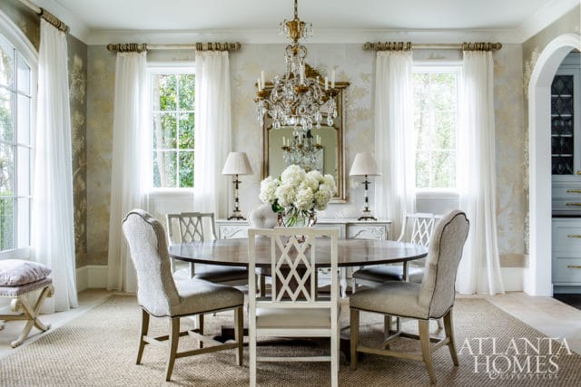 Dining room with oval wood table surrounded by upholstered chairs, ornate crystal chandelier, and patterned wallpaper.