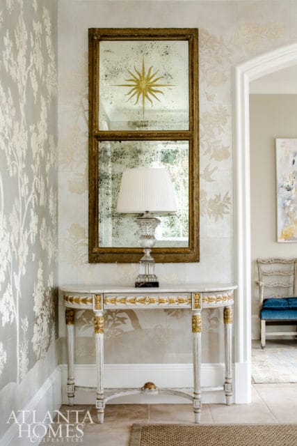 Hallway console table with marble top and gold accents, topped with a crystal lamp beneath a distressed mirror with a sunburst design.