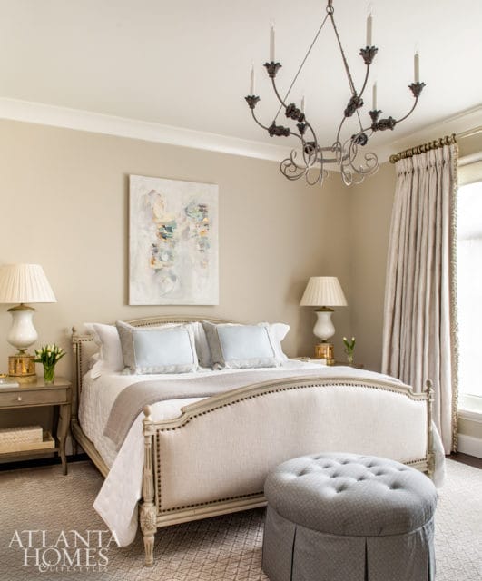 Neutral bedroom with a tufted upholstered bed, light blue accent pillows, a grey ottoman, and a wrought iron chandelier.