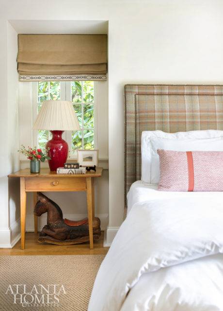 A guest bedroom reveals a menswear attitude with a plaid upholstered headboard and simple wooden table with roman shades. Stronger color comes from a pillow in a Fermoie fabric through Jerry Pair.