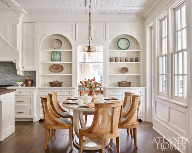 White kitchen with arched built-in shelves displaying decorative plates and baskets, a round dining table set for four, and cane-backed chairs.