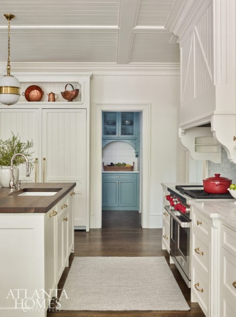 White kitchen with a coffered ceiling, a large island with a dark wood countertop and sink, and a doorway leading to a blue pantry with arched shelving.