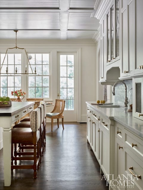 White kitchen with coffered ceiling, brass hardware, marble countertops, and a large island with leather and patterned stools.