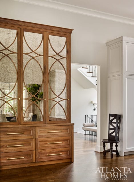 Wooden built-in cabinet with mirrored doors featuring an ornate geometric pattern, six drawers with brass hardware, and a view of a staircase and seating area in the background.
