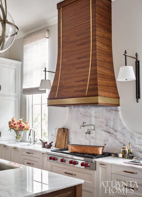 Cream kitchen with a large wooden range hood accented with brass trim, marble backsplash, and a professional gas range with red knobs.