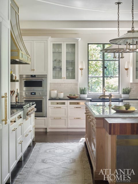 White kitchen with brass hardware, double ovens, glass-front cabinets, and a large window overlooking greenery. A patterned runner rug is on the dark wood floor.