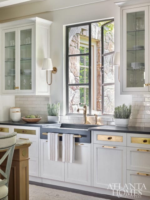 White kitchen with a dark apron sink, brass faucet, and gold hardware on white cabinets. A large window with a black metal frame looks out onto a stone exterior.