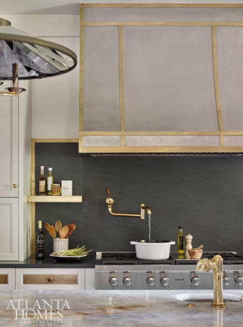 Modern kitchen with dark slate backsplash, brass pot filler faucet, and stainless steel Wolf range. White pot on stove with water running from faucet.