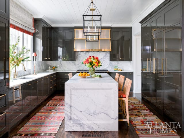 Dark gray high-gloss kitchen with marble island, brass hardware, woven bar stools, and colorful runner rugs.