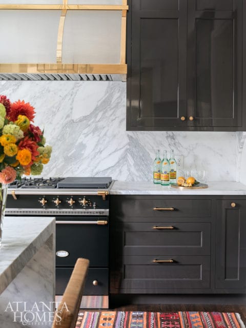 Dark gray kitchen cabinets with brass hardware, marble backsplash and countertops, and a black range with brass accents.