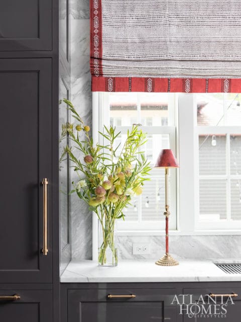 Dark gray kitchen cabinets with brass hardware, white marble backsplash, and a patterned Roman shade above a window. A vase of greenery and a red candlestick lamp sit on the counter.